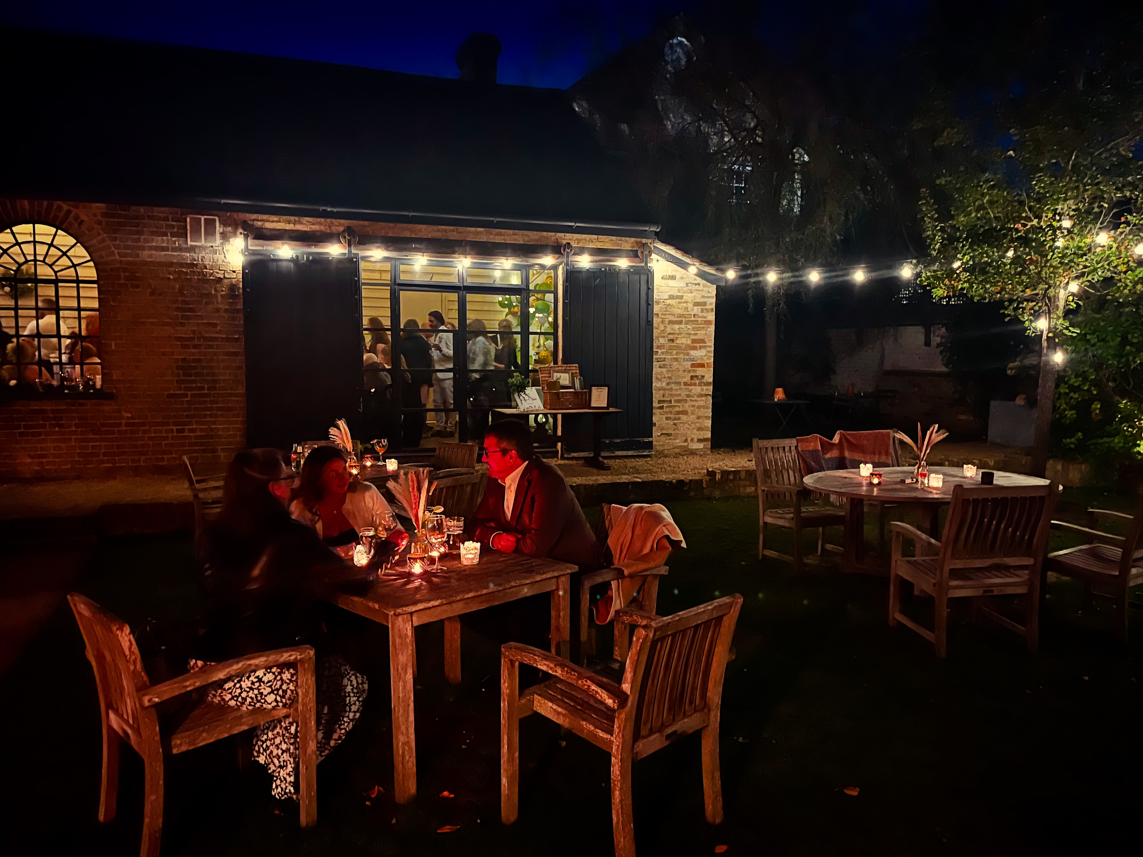 Guests dining in the garden at night by candlelight and festoon lights